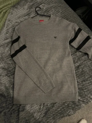 Grå stickad tröja från Guess - Snygg grå stickad tröja från Guess med svarta ränder på ärmarna och metallfärgad logga på bröstet. Tröjan har rund halsringning och ribbade muddar vid ärmslut och nederkant. Perfekt för dig som gillar stilrena och sportiga detaljer.