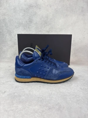 Blå sneakers från Valentino - Snygga blå sneakers från Valentino med ikoniska nitar på sidan. Skorna har en mix av mocka och läder, blå snörning och en kontrasterande beige sula. Perfekta för dig som vill ha en unik och trendig look.