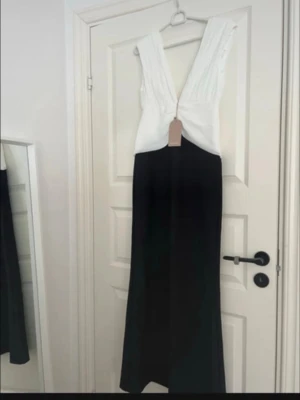 Svartvit långklänning från Meshki - Elegant långklänning från Meshki i svart och vitt. Klänningen har djup v-ringning, breda axelband med veckade detaljer och en figurnära passform upptill. Kjoldelen är svart och rak. Perfekt för dig som vill ha en stilren och modern look. Prislappen kvar!