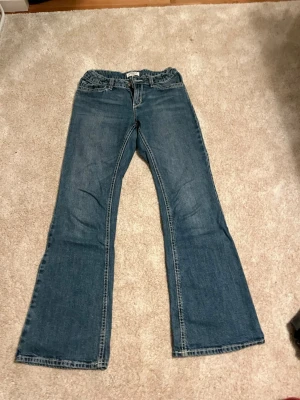 Blå bootcut jeans från Lindex - Säljer ett par blå bootcut jeans från Lindex, modell Freja. Jeansen har klassisk femficksdesign, dragkedja och knapp framtill samt kontrastsömmar. Tillverkade i slitstark denim med lätt tvättad look. Perfekta för en avslappnad och trendig stil.