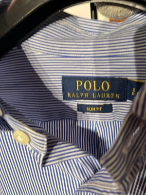 Blårandig skjorta från Polo Ralph Lauren - Klassisk blårandig skjorta från Polo Ralph Lauren med button-down krage och broderad logga på bröstet. Slim fit-modell med långa ärmar och vita knappar. Tillverkad i mjuk bomull som känns skön mot huden.