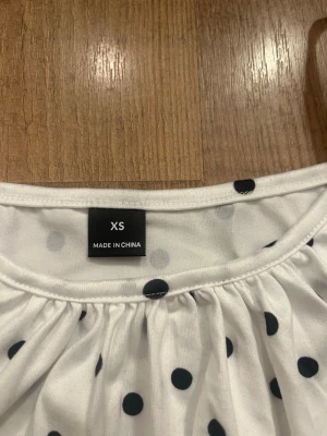 Vit prickig linnetopp med smala axelband - Söt vit linnetopp med svarta prickar och smala axelband. Toppen har en lös passform och är perfekt för varma dagar. Materialet känns mjukt och lätt mot huden. Enkel och stilren design som passar till många olika outfits.