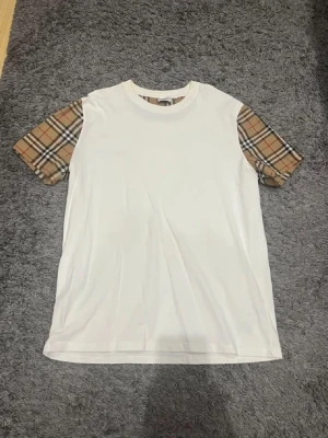 Vit Burberry t-shirt med rutiga ärmar. Helt oanvänd. Storlek L men sitter som M - Säljer en clean vit t-shirt från Burberry med deras ikoniska beigea, bruna och svarta rutiga mönster på ärmarna och insidan av kragen. Klassisk passform och rund hals. 