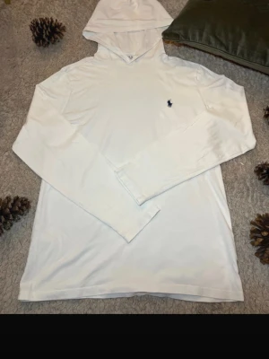 Vit hoodie från Polo Ralph Lauren - Snygg vit hoodie från Polo Ralph Lauren med klassisk broderad logga i mörkblått på bröstet. Hoodien har långa ärmar, huva och är gjord i mjukt bomullstyg. Perfekt för en clean och stilren look. Det är en lapp som saknas.