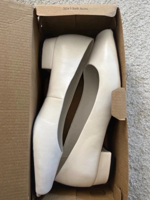 Vita ballerinaskor med låg klack MNG - Stilrena vita ballerinaskor från MNG med fyrkantig tå och låg, bred klack. Skorna är tillverkade i ett slätt material som ger en modern och minimalistisk look. Perfekta för dig som gillar enkel och trendig design.