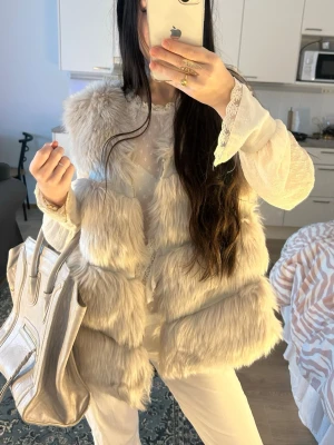 Ljusgrå fluffig pälsväst från Miss Look - Supersnygg ljusgrå pälsväst från Miss Look med fluffig fuskpäls och mjukt foder. Västen har en voluminös look och är perfekt för att lyfta vilken outfit som helst. Stängs framtill och har en modern, oversized känsla.