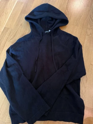 Svart hoodie med dragsko - En enkel svart hoodie med huva och dragsko. Hoodien har långa ärmar och ribbade muddar vid ärmslut och nederkant. Perfekt för en avslappnad stil och passar till det mesta. Materialet ser ut att vara mjukt och bekvämt.