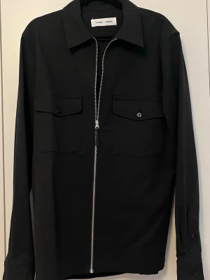 Svart overshirt - Samsøe Samsøe - Stilren svart overshirt från Sandro Paris med rak passform, två bröstfickor med lock och knapp samt hel dragkedja framtill. Klassisk krage och långa ärmar. Perfekt lager på lager-plagg i ett mjukt tyg.