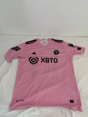 Rosa Inter Miami matchtröja Messi - Snygg rosa Inter Miami CF matchtröja från Adidas med svarta detaljer och tryck. Messi och nummer 10 på ryggen. Tröjan har klubbmärke på bröstet, sponsorloggor och klassiska Adidas-ränder på axlarna. Perfekt för fotbollsfans som vill sticka ut.