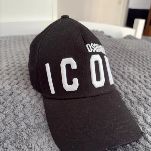 Svart Dsquared2 ICON keps - Säljer en svart keps från Dsquared2 äkta med stor vit brodyrtext 'ICON' framtill och mindre Dsquared2-logga ovanför. Klassisk böjd skärm och justerbar baksida. Tillverkad i bomull för en bekväm och snygg look.