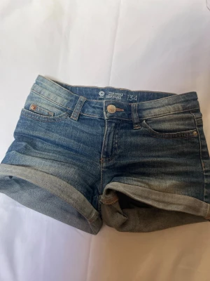 Blå denimshorts med upprullad kant - Säljer ett par klassiska blå jeansshorts från Your Denim med upprullad kant och femficksmodell. Shortsen har normal passform och stängs med knapp och dragkedja. 