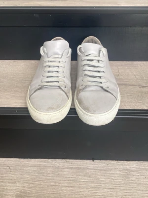 Gråa låga sneakers i läder - Stilrena grå sneakers med lågt skaft och klassisk siluett. Skorna har vita snören och sula, samt är tillverkade i slätt läder för en clean look. Passar perfekt till jeans eller chinos för en avslappnad vibe.