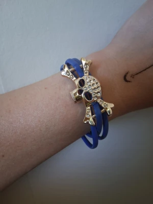 Blått armband med dödskalle - Snyggt blått armband med en stor guldfärgad dödskalle och korslagda ben dekorerade med små glittriga stenar. Armbandet har flera band som ger en cool och edgy look. Perfekt för dig som gillar statement-smycken och vill sticka ut.