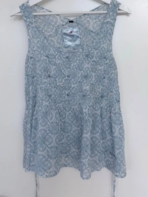 Fin knytblus från Bondelid! I bomull och viskos. - Ärmlös blus med mönster i blått och vitt. Broderade detaljer. Knytband i sidorna. En blandning av bomull och viskos.  L 63 cm   