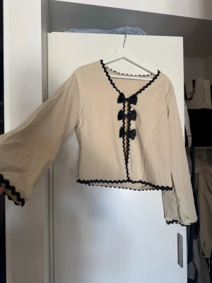 Beige blus med svarta rosetter - Trendig beige blus med långa, vida ärmar och dekorativa svarta rosetter framtill. Svart vågig kant längs halsringning, ärmslut och nederkant ger en unik look. Perfekt för dig som vill sticka ut med en söt och stilren topp. Aldrig använd 