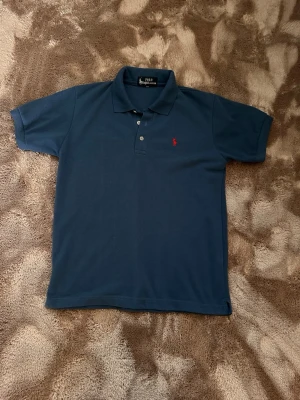 Ralph louren pike - Klassisk blå pikétröja från Polo Ralph Lauren med korta ärmar och krage. Tröjan har två knappar framtill och den ikoniska röda logotypen broderad på bröstet. Tillverkad i mjuk bomull som känns skön mot huden.