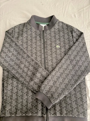 Lacoste zip-up track jacket  - Säljer en stilren och äkta Lacoste ziptröja i mörkgrå färg med exklusivt geometriskt monogrammönster över hela plagget. Broderad Lacoste-krokodil på bröstet och hög kvalitet i både material och detaljer.  ✔ 100 % äkta Lacoste ✔ Genomgående monogrammönster ✔ Dragkedja framtill ✔ Ribbad krage, ärmslut och nederkant ✔ Bekväm passform ✔ Perfekt för både vardag och streetwear  Tröjan är i fint skick och ger en exklusiv look som sticker ut utan att vara för prålig.