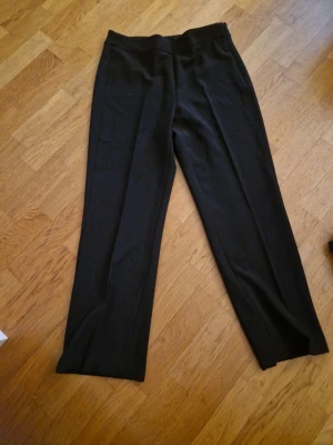 Svarta kostymbyxor med pressveck - Snygga svarta kostymbyxor med klassiskt pressveck och rak passform. Byxorna har dold dragkedja och hake i midjan för en clean look. Perfekta till skjorta eller stickad tröja. Materialet känns mjukt och följsamt. Haken behöver eventuellt sys fast, men den går att använda.