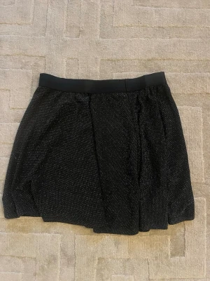 Svart glittrig kjol med resårmidja - Kort svart kjol med diskret glittrigt tyg och bred resår i midjan. Kjolen har en lätt utställd form och är perfekt för att skapa en trendig look. Materialet är mjukt och följsamt, vilket gör den bekväm att bära hela dagen.