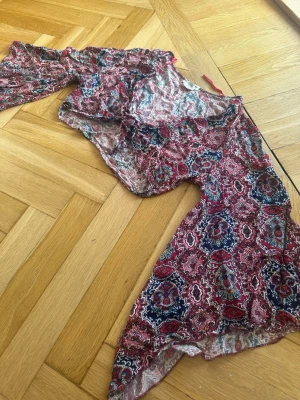 Boho topp med knytning - Fin boho-inspirerad topp med vackert paisleymönster i vinröda, rosa och marinblå toner köpt i Grekland. Modellen knyts framtill och har en feminin passform som framhäver dekolletaget på ett smickrande sätt. De luftiga ärmarna ger en romantisk och avslappnad känsla.  Perfekt till jeans, shorts eller kjol under sommaren, på semestern eller till festivaler. 🌸✨  Storlek M men passade mig som brukar ha XS-S