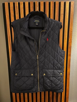 Marinblå quiltad dunväst Polo Ralph Lauren - Snygg marinblå dunväst från Polo Ralph Lauren med quiltat mönster, dragkedja framtill och två stora fickor med tryckknappar. Klassisk röd logga broderad på bröstet och hög krage för extra stilpoäng. Perfekt lager på lager-plagg för kyliga dagar. Den har ett brännmärke vid sidan av loggan, men det går säkert att laga lätt!