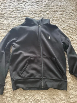 Svart hoodie från Polo Ralph Lauren - Säljer en svart hoodie från Polo Ralph Lauren med dragkedja framtill och klassisk logga broderad på bröstet. Hoodien har känguruficka och justerbar huva med snören. Tillverkad i mjuk bomullsblandning, perfekt för en avslappnad stil.