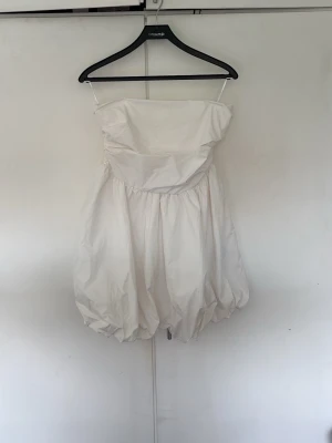 Vit axelbandslös kort klänning - Säljer en vit axelbandslös kort klänning med ballongformad kjol och smock i ryggen. Klänningen har en luftig och voluminös siluett, perfekt för sommarens alla tillfällen. Materialet känns lätt och bekvämt mot huden.