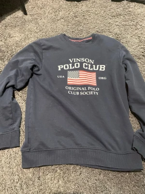 Blå sweatshirt från Vinson Polo Club - Mörkblå sweatshirt från Vinson Polo Club med tryck av amerikanska flaggan och text på bröstet. Tröjan har rund halsringning, ribbade muddar vid ärmslut och nederkant. Perfekt för en avslappnad och sportig stil.