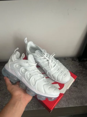 Nike Air Vapormax Plus vita sneakers - Nike Air Vapormax Plus sneakers i helvitt med futuristisk design och genomskinlig sula. Ovandelen är i textil och syntet med tydliga detaljer och snörning. Perfekta för dig som vill ha en clean och modern look.
