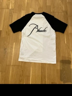 Svartvit t-shirt från Rhude - Snygg svartvit t-shirt från Rhude med kontrasterande svarta raglanärmar och vit kropp. Stort Rhude-tryck på ryggen och mindre logga framtill. Perfekt för en stilren och sportig look.