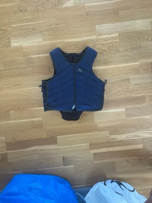 Mörkblå skyddsväst från CRW - Säljer en mörkblå skyddsväst från riders company med dragkedja framtill och justerbara sidor. Västen har vadderade paneler för extra skydd och en sportig look. Perfekt för ridning eller andra aktiviteter där extra skydd behövs.