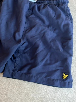 Marinblå shorts från Lyle & Scott - Snygga marinblå shorts från Lyle & Scott med elastisk midja och den klassiska gula loggan broderad vid benet. Perfekta för sommardagar och enkel att matcha med olika toppar.
