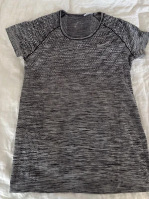 Grå Nike Dri-Fit träningströja - Säljer en gråmelerad Nike Dri-Fit träningströja med korta ärmar och rund hals. Materialet är lätt och andas, perfekt för intensiva träningspass. Diskret Nike-logga på bröstet och snygg sportig design. Passar dig som vill ha funktion och stil på gymmet.