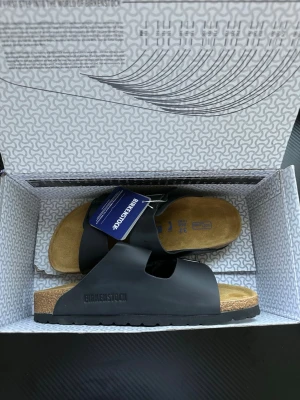 Svarta Birkenstock Arizona sandaler - Helt nya Klassiska Birkenstock Arizona sandaler i svart med två justerbara remmar och metallspännen. Fotbädden är mjuk och tillverkad i mocka och kork för extra komfort. Yttersulan är i EVA och ovandelen i syntetiskt material. Perfekta för en avslappnad och trendig look.