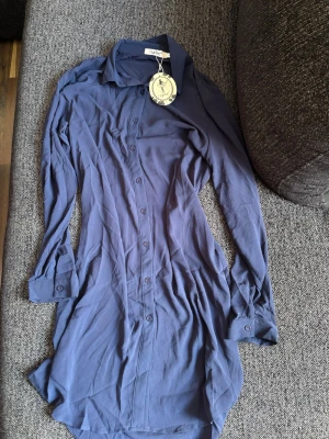 Blå skjortklänning från NÜMPH - Snygg blå skjortklänning från NÜMPH med klassisk krage och knappar hela vägen framtill. Långärmad modell i lätt och mjukt material, perfekt för en avslappnad men stilren look.