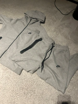 Grå Nike mjukisdress med huva - Säljer en grå mjukisdress från Nike med dragkedja och huva på tröjan samt matchande byxor med snörning i midjan. Båda delarna har Nike-logga och tröjan har svarta detaljer vid fickorna. Perfekt för chill dagar eller träning.