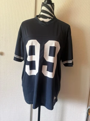 Mörkblå oversized sporttopp med 99 - Cool mörkblå sporttopp med stora vita siffror 99 framtill och texten ROCKET på ryggen. Oversized passform och korta ärmar med vita ränder. Perfekt för en avslappnad och sportig look.
