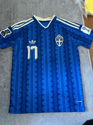 Blå Sverige retro fotbollströja Adidas - Retroinspirerad blå fotbollströja från Adidas med Sveriges landslagsemblem och nummer 17 på bröstet. Tröjan har klassiska vita Adidas-ränder på axlarna, v-ringad krage och detaljer i gult. Tillverkad i lätt och ventilerande material, perfekt för match eller träning.