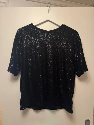 Svart paljettopp från H&M - Säljer en svart topp från H&M täckt med glittriga paljetter. Toppen har rund halsringning och korta ärmar. Toppen har en knapp i nacken för enkel påklädning.