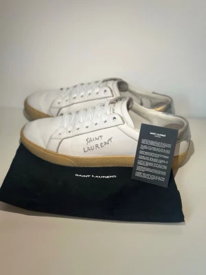 Saint Laurent vita sneakers i läder - Säljer ett par vita Saint Laurent sneakers i läder med silverdetaljer på hälen och handskriven logga på sidan. Skorna har klassisk låg profil, vita snören och beige gummisula. Kommer med originalpåse och äkthetskort.
