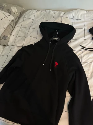 Svart hoodie från Ami Paris med röd logga - Svart hoodie från Ami Paris med klassisk huva och dragsnören. På bröstet sitter Ami Paris ikoniska röda hjärta-logga. Hoodien har en avslappnad passform och är tillverkad i mjuk bomull för maximal komfort.