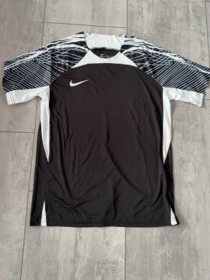 Svart och vit Nike träningströja - Snygg svart och vit träningströja från Nike med korta ärmar och grafiskt mönster på axlarna. Tillverkad i ett lätt och ventilerande material som passar perfekt till sport och träning. Klassisk Nike-logga på bröstet.