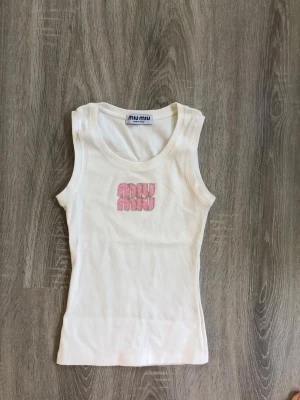 Vit linnetopp från Miu Miu - Snygg vit linnetopp från Miu Miu med broderad rosa logga framtill. Toppen är ärmlös och har en klassisk rund halsringning. Perfekt att styla till jeans eller kjol för en trendig look.