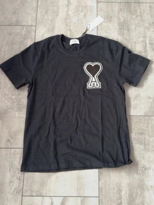 Svart t-shirt från Ami Paris - Snygg svart t-shirt från Ami Paris med det ikoniska hjärta-A broderiet på bröstet. Klassisk passform och rund halsringning. Tillverkad i mjuk bomull som känns skön mot huden. Perfekt för dig som gillar stilrena plagg med en twist.