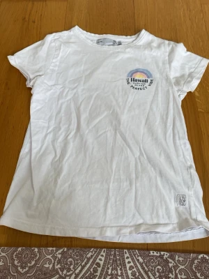 Vit Hawaii t-shirt med regnbågstryck - Vit t-shirt från Name It med färgglatt regnbågstryck och texten 'Hawaii Surfing Island The Perfect Vibe' både fram och bak. Klassisk rund hals och korta ärmar. Tillverkad i mjuk bomull, perfekt för en avslappnad och cool stil.