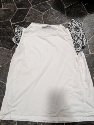 Vit topp med svarta spetsdetaljer stl xl - Säljer en vit topp med breda, svarta spetsärmar i blommigt mönster. Toppen har rund halsringning och ärmlös design med dekorativa spetsar som ger en unik look. Perfekt för dig som vill ha något extra till outfiten. Skulle säga att stl motsvarar 42