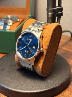 Citizen klocka med blå urtavla - Stilren klocka från Citizen med blå urtavla, silverfärgade siffror och datumvisning. Armbandet är i blankt metall och boetten har en klassisk rund form. Perfekt för dig som gillar en clean och tidlös look.