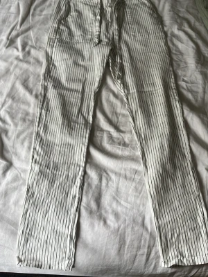 Vita linnebyxor med svarta ränder - Säljer ett par vita linnebyxor med tunna svarta vertikala ränder och dragsko i midjan. Byxorna har raka ben och avslappnad passform, perfekta för en chill och stilren look. Materialet känns lätt och luftigt, perfekt för varma dagar. Finns ingen lapp å har glömt vilken storlek men tror 34