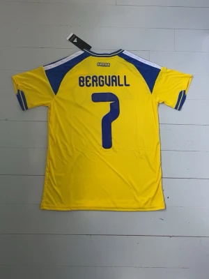 Sverige matchtröja Adidas | Bergvall - Gul och blå matchtröja från Adidas för Sveriges landslag med tryck 'Bergvall' och nummer 7. Tröjan har korta ärmar, blå detaljer på axlar och ärmslut samt broderad landslagssköld på bröstet. Tillverkad i lätt och ventilerande material, perfekt för fotboll.