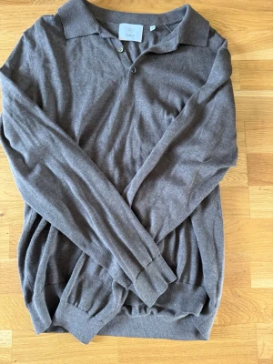 Brun långärmad tröja från Bläck - Snygg brun långärmad tröja från Bläck med krage och knappar vid halsen. Tröjan är tillverkad i ett mjukt, tunt stickat material och har ribbade muddar vid ärmslut och nederkant. Perfekt för en stilren och avslappnad look.